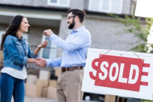 Real estate agent giving woman keys in front of a home with a sold sign in the yard. 