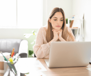 Young white woman with long brown hair staring a  laptop sadly.